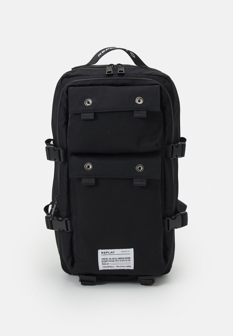 Sac à dos noir avec deux poches à rabat à l'avant munies d'œillets métalliques, des boucles latérales, une poignée supérieure avec texte et une étiquette blanche en bas.