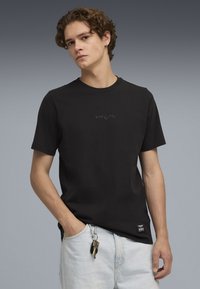Camiseta de algodón negra con cuello redondo, que presenta un sutil logo en relieve en el pecho y una pequeña etiqueta en el dobladillo, combinada con pantalones cortos de mezclilla claros.