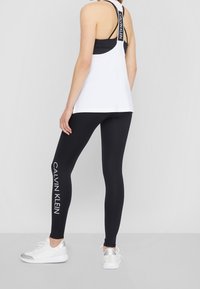 Bílý sportovní top s designem na zádech, vrstvený přes černou podprsenku, doplněný černými legínami s nápisem "CALVIN KLEIN" v bílé barvě.