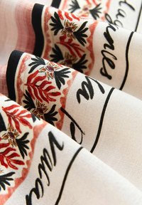 Tissu avec fond beige, motif floral rouge et noir, bords bordés de noir, et texte cursif noir partiellement visible.