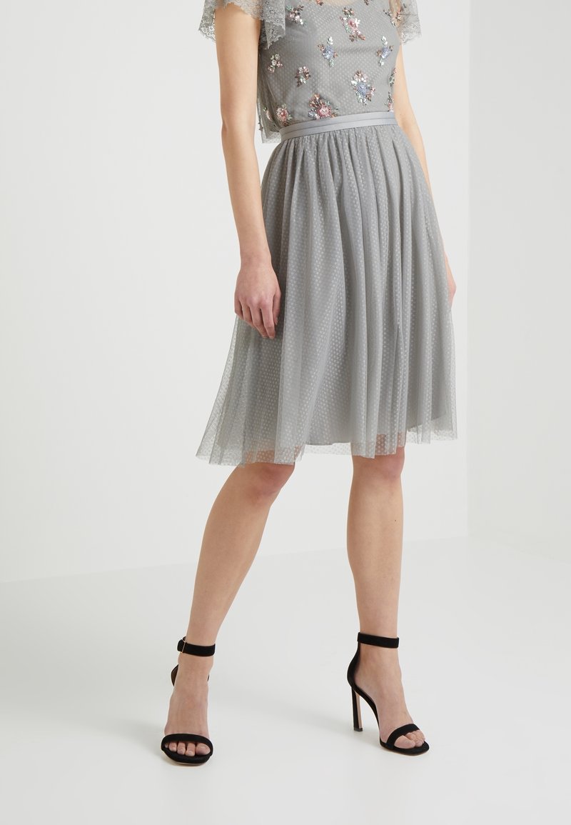 Femme portant une jupe en tulle grise jusqu'aux genoux et un haut brodé floral, associée à des escarpins noirs à bride cheville, debout sur un sol blanc.