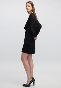 Schwarzes Kleid mit langen, fließenden Ärmeln, verziert mit kreisförmigen Perlenmustern. Kombiniert mit schwarzen High Heels. Sanfte Stofftextur.