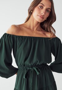Dunkelgrünes, schulterfreies Kleid aus fließendem Stoff mit elastischem Bund und gebundenem Detail, mit langen, lockeren Ärmeln und einem gestuften Design.