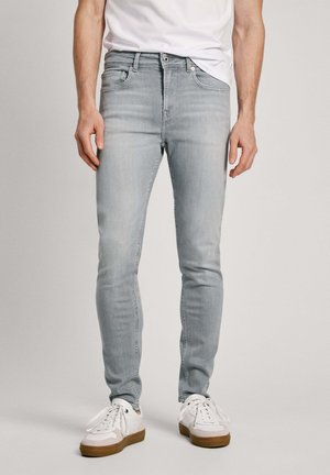 Muž nosící světle šedé úzké džíny, bílé tenisky s gumovou podrážkou a bílé tričko, stojící před jednobarevným pozadím.