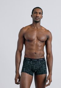 Μαύρα σλιπ μποξέρ με ελαστική μέση με λογότυπο, με επαναλαμβανόμενο μοτίβο λογότυπου. Το μοντέλο στέκεται όρθιο με ουδέτερη έκφραση.