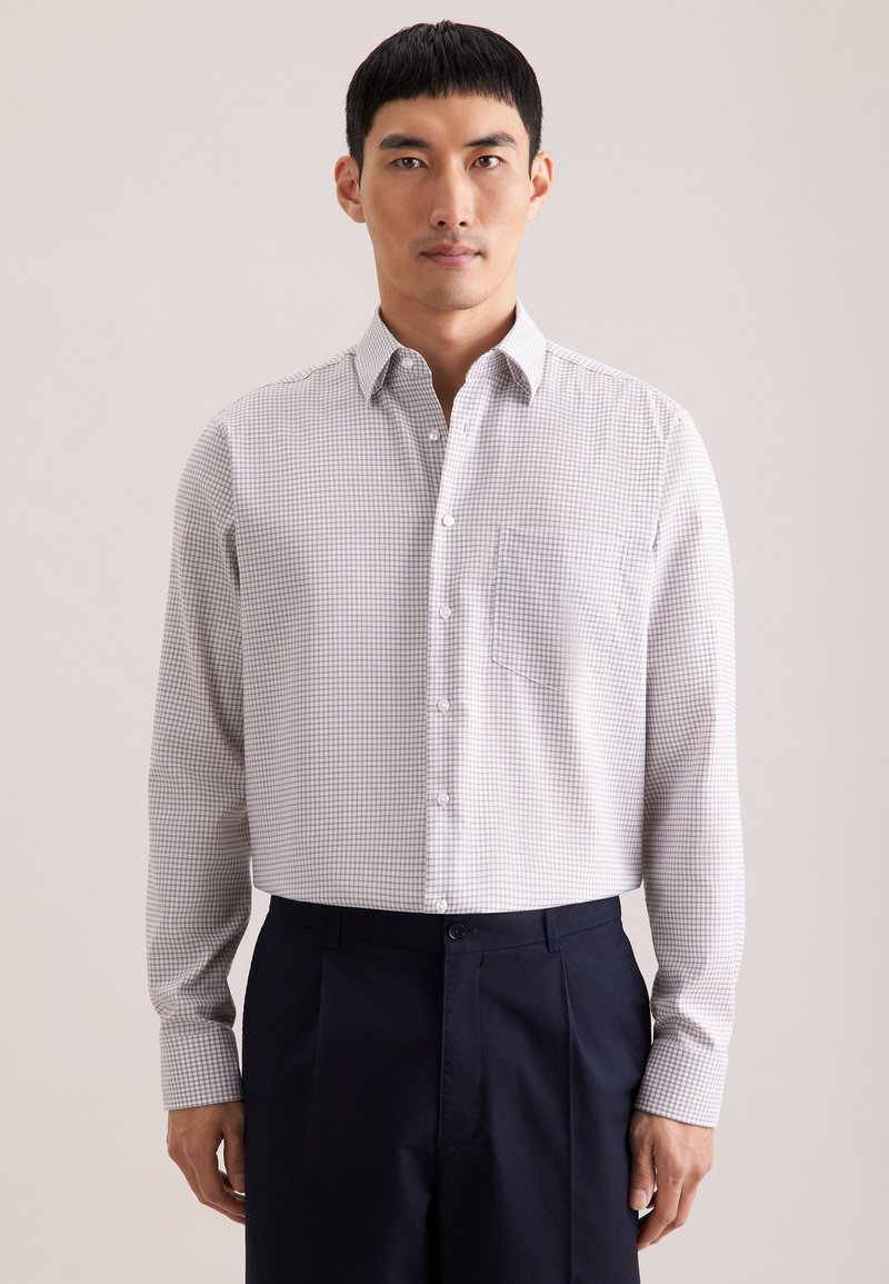 Biała koszula z długim rękawem w drobny brązowy wzór w kratkę, z kołnierzykiem na guziki, pojedynczą kieszenią na piersi, noszona z granatowymi spodniami.