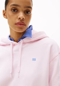 Felpa leggera rosa con un logo ricamato blu, dotata di cappuccio con stringa di regolazione. Sotto, è visibile una camicia colorata a righe blu e bianche con colletto.