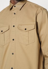 Camisa beige de botones con dos bolsillos en el pecho, uno de los cuales está bordado con "HUGO" en hilo claro.