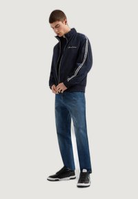 Chaqueta de bombardeo azul marino con franjas laterales blancas, cierre con cremallera, puños y dobladillo elásticos, combinada con jeans azules y zapatillas negras.