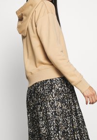 Beige hoodie van zacht materiaal, met ribgebreide boorden en onderrand, gecombineerd met een geplooide zwarte rok met een kleurrijk abstract patroon.