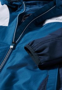 Modrá a tmavě modrá větruodolná bunda s zipovým zapínáním, elastickými manžetami a kapucí. Texturovaná tkanina s barevnými blokovými prvky.