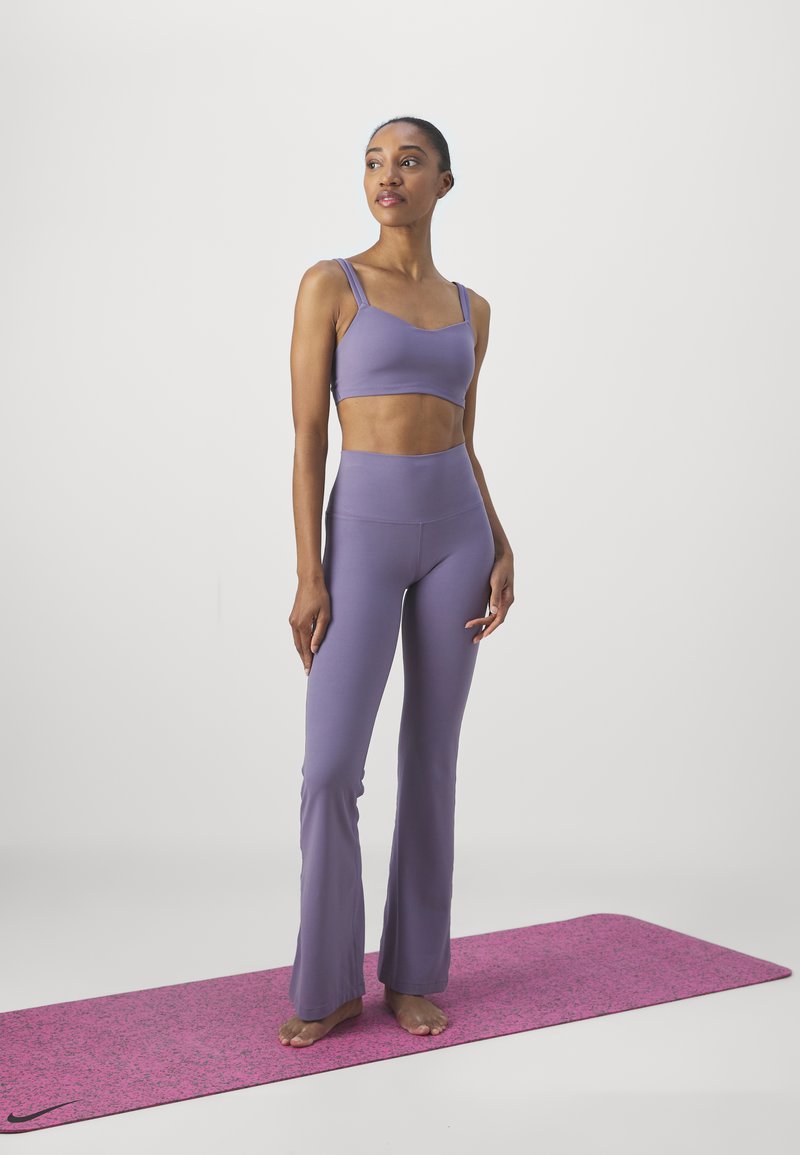 Top corto de lavanda y leggings acampanados hechos de un tejido suave, acompañados de una esterilla de yoga de color rosa con manchas. Modelo de pie descalzo.