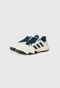 Een paar crèmekleurige sportschoenen met zwarte strepen, donkergroene veters en zwarte zolen op een witte achtergrond.