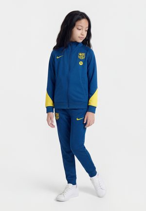 Dziecko stoi, ubrane w niebiesko-żółty dres FC Barcelona Nike z białymi tenisówkami, patrzy w dół i na bok.