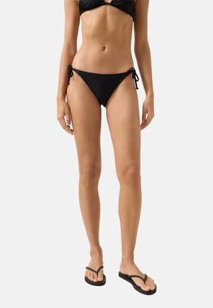 Frau trägt einen schwarzen String-Bikini mit seitlichen Bindebändern und schwarzen Flip-Flop-Sandalen, stehend vor einem schlichten hellen Hintergrund.