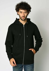 Felpa nera con zip realizzata in tessuto morbido, con tasca frontale, cappuccio regolabile con lacci e polsini a coste. Indossata con jeans blu casual.