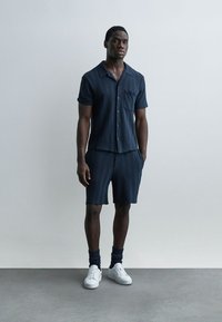 Homme debout contre un mur gris clair portant une chemise à manches courtes bleu marine à boutons, un short assorti, des chaussettes foncées et des baskets blanches.