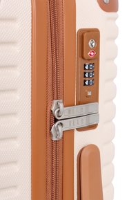 Hard-shell suitcase in beige with brown zipper and accents. Features dual zippers, combination lock, and textured surface with vertical lines.