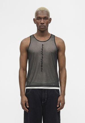 Ung mand med kort blondt hår iført en gennemsigtig sort tanktop over en hvid undertrøje og løstsiddende mørke bukser, stående mod en ensfarvet hvid baggrund.