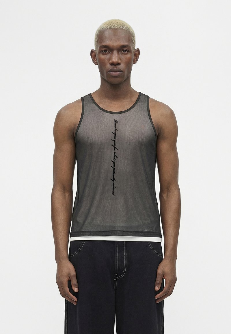 Νέος άνδρας με κοντά ξανθά μαλλιά φοράει διάφανο μαύρο φανελάκι πάνω από λευκή μπλούζα και φαρδιά σκούρα παντελόνια, στέκεται μπροστά σε απλό λευκό φόντο.