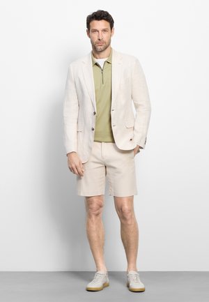 Man draagt een beige blazer en korte broek met lichtgroene zip-up polo en witte sneakers, staand tegen een witte achtergrond.