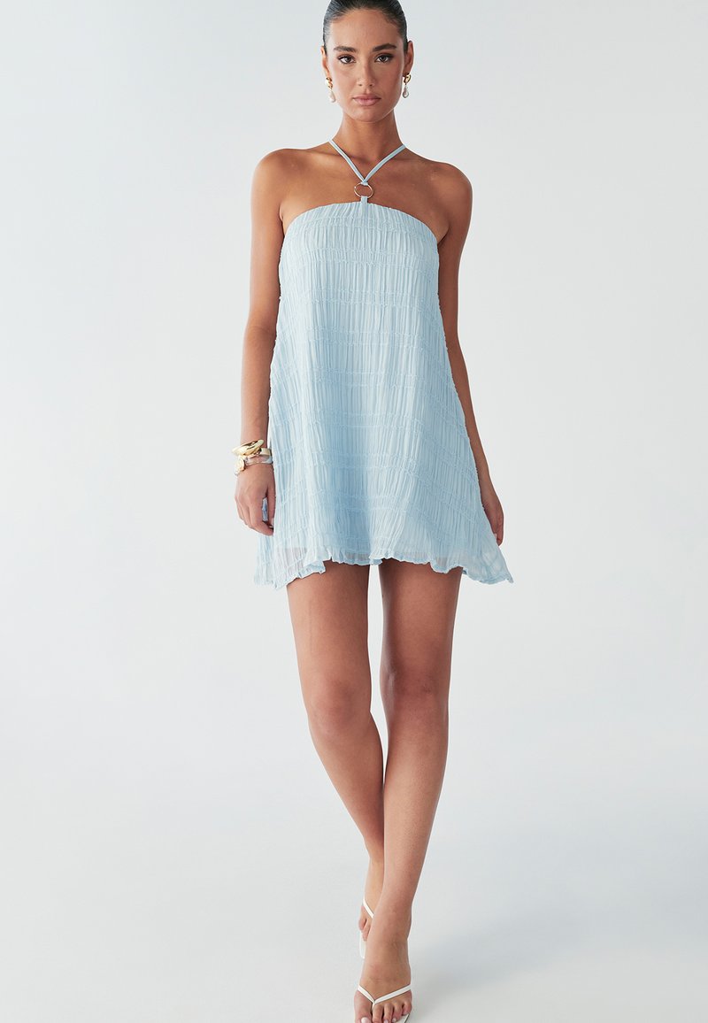 Een model staat in een lichtblauwe strapless jurk met een gegatherde textuur, draagt gouden armbanden en strappy witte sandalen, tegen een eenvoudige achtergrond.
