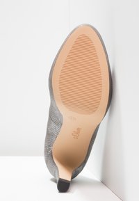 Chaussure à talons hauts texturée grise inclinée vers le haut, montrant une semelle beige lisse avec un motif de grip léger et le nom de la marque "s.Oliver" estampillé.