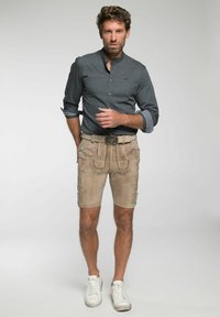 Beige Lederhosen mit dekorativen Nähten, kombiniert mit einem dunkel gemusterten Langarmshirt und weißen Turnschuhen.