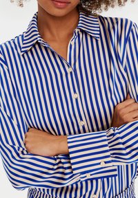 Blå och gräddfärgad randig skjorta med knappar och krage, med vertikala ränder, en mjuk textur och knappar på ärmsluten.