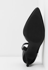 Escarpin noir à bout pointu avec semelle texturée et bride à la cheville, positionné droit contre un fond d’angle blanc.