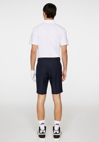 Vit polo-tröja, marinblå shorts och vita strumpor. Observerad bakifrån, vilket visar en avslappnad passform och släta texturer. Svarta sportskor.