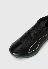 Scarpa da ginnastica nera con superficie testurizzata, suola verde e accento giallo-verde di lato. Presenta un design con lacci e una parte superiore traspirante.