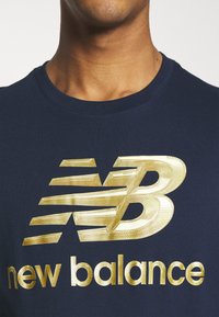 Marinblå T-shirt med en framträdande guld New Balance-logotyp. Designen har texturerade detaljer och en rundad halsringning.