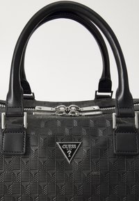 Bolso de mano negro con un patrón texturizado, herrajes de plata, doble cremallera y un logotipo triangular destacado en la parte frontal. Asas de cuero suave.