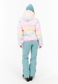 Puff jacket im pastellfarbenen Batikmuster mit Kapuze, kombiniert mit hellblauen Skihosen und schwarzen Schneestiefeln.