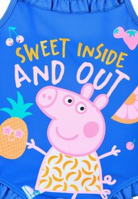 Blauer Badeanzug mit einer Grafik eines rosa Schweinchen-Charakters, mit dem Text "SÜSS INNEN UND AUßEN" und bunten Obstdesigns, darunter eine Ananas und Kirschen.