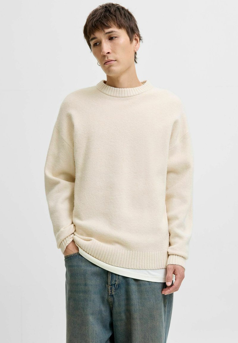 Cream sweater with a ribbed neckline, drop shoulders, and a soft texture. Paired with loose denim jeans in a faded blue tone.
