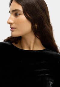 Schwarzes Samtoberteil mit runder Ausschnitt. Modell trägt einen goldenen Creolenohrring. Glattes Haar, das über die Schulter fällt. Neutraler Hintergrund.