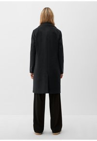 Svart ullkappa med krage, rak design, medellång, minimal sömnad och knappdekorationer på ärmarna, bärs över vida svarta byxor.