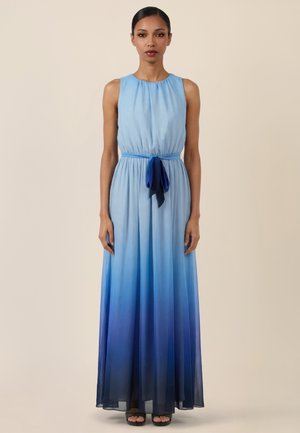 Frau trägt ärmelloses Maxi-Kleid in Blau-Ombré mit Taillengürtel, steht vor schlichtem beigem Hintergrund.