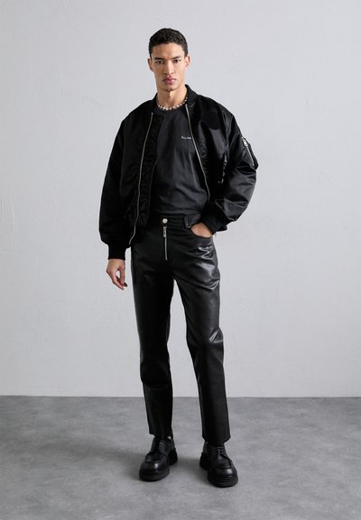 Veste bomber noire, chemise noire ajustée et pantalon en cuir noir. Détails notables de fermeture éclair en argent sur le pantalon et chaussures noires massives.