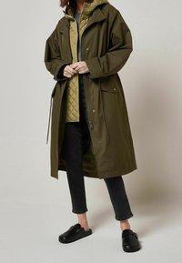 Manteau imperméable vert olive avec une taille cintrée, de grandes poches et une capuche, superposé à une veste matelassée olive, associé à un jean noir et des chaussures à enfiler.