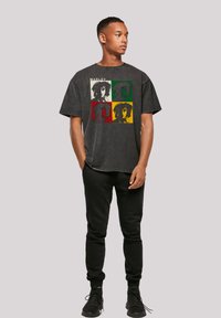 Schwarzes T-Shirt mit kurzen Ärmeln, das eine Grafik von vier stilisierten Porträts mit roten, grünen und gelben Akzenten zeigt. Kombiniert mit schwarzen Sweatpants und Schuhen.