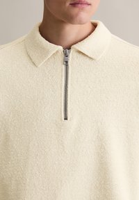 Jasno beżowy zapinany na zamek polar o miękkiej, teksturowanej tkaninie, z wyraźnym kołnierzem i srebrnym zamkiem. Projekt ma swobodny, dopasowany krój.