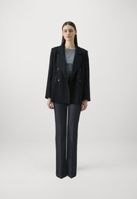 Blazer nero doppiopetto sopra una maglietta grigia, abbinato a pantaloni in denim scuro dritti con cuciture chiare. Accento di una semplice collana d'oro.