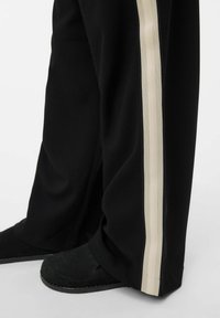 Svarta vida byxor i mjukt tyg med en krämfärgad sidrand. Bensluten slutar vid vristen och de är kombinerade med svarta tofflor.