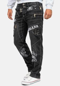 Jeans cargo in denim nero con numerose tasche, accessori in argento, ricami bianchi e un taglio ampio. Dotati di zip e borchie decorative.