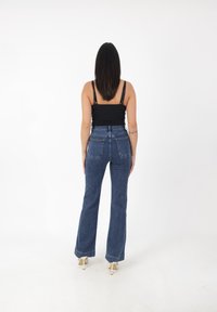 Jeans z wysokim stanem, rozszerzane, w ciemnoniebieskim deninie, z pięcioma kieszeniami i widocznymi szwami. Modelka w jasnych szpilkach.