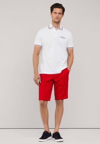 Camisa polo blanca con cuello a rayas rojas y azules, logo en el pecho, combinada con pantalones cortos rojos y zapatos oscuros, sobre un fondo neutro.