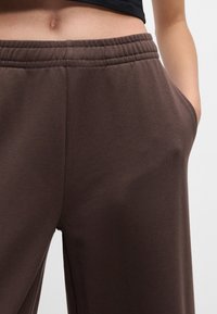 Καφέ φόρμες με ελαστική μέση, πλαϊνές τσέπες και λεία υφή, εμφανιζόμενες από τη μέση και κάτω. Λεπτομέρειες ραφής κοντά στη μέση.